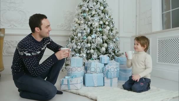 Padre feliz con su hijo sentado cerca del árbol de Navidad y bebiendo té caliente — Vídeo de stock