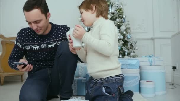 Ocupado padre hablando por teléfono mientras su hijo bebe cacao cerca del árbol de Navidad — Vídeo de stock