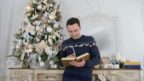 Fiatal férfi kötött pulóver szarvast olvasó egy könyv mellett a karácsonyfa — Stock videók