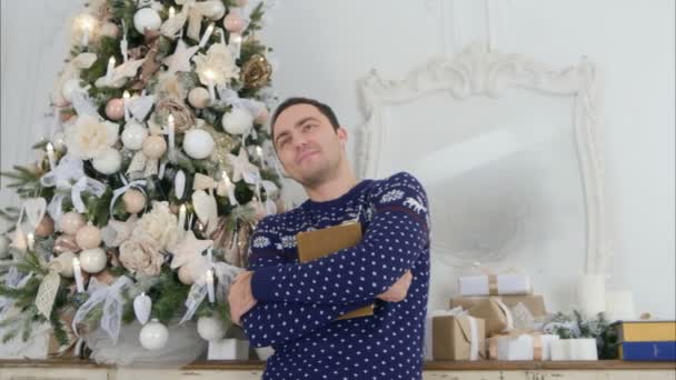 Ung mand holder en bog til brystet og står drømmende ved siden af juletræet – Stock-video