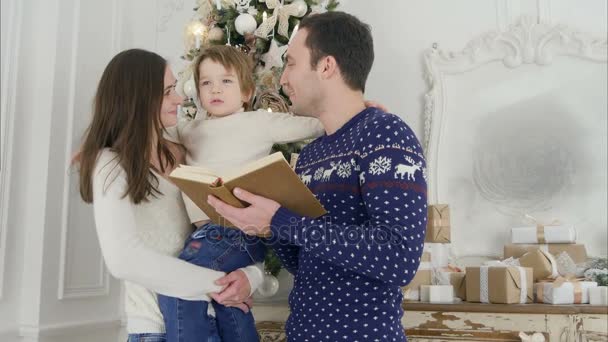 Šťastný otec čtení Vánoční příběh zatímco matka drží jejich veselá syn před vánoční stromeček — Stock video