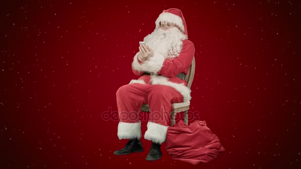 Weihnachtsmann lacht laut, als er auf seinem Handy auf rotem Hintergrund mit Schnee spricht — Stockvideo