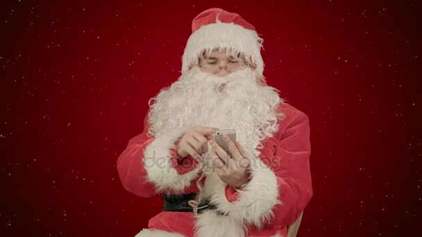 Santa claus να διαβάζει και να στέλνει μηνύματα κειμένου από το τηλέφωνο κυττάρων σε κόκκινο φόντο με χιόνι — Αρχείο Βίντεο