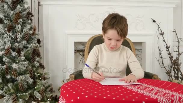 Menino sério escrevendo carta para o Papai Noel sentado na mesa perto da árvore de Natal — Vídeo de Stock