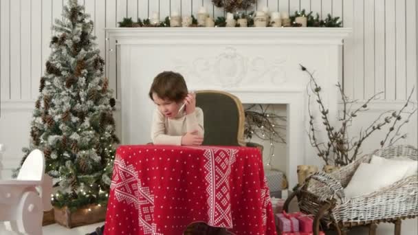 Cute little boy calling Santa while sitting on a big armchair at home over Chirstmas tree background — Stock Video