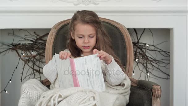 Linda menina embrulhada em cobertor de Natal branco lendo um livro sentado em uma poltrona perto da lareira decorada — Vídeo de Stock