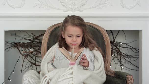 Preciosa niña envuelta en manta blanca de Navidad explorando un libro sentado en un sillón cerca de la chimenea decorada — Vídeos de Stock