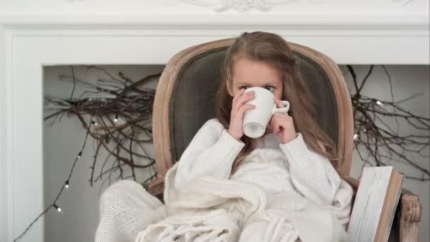 Bambina seduta su un'accogliente sedia avvolta in una coperta che beve tè vicino al camino di Natale — Video Stock