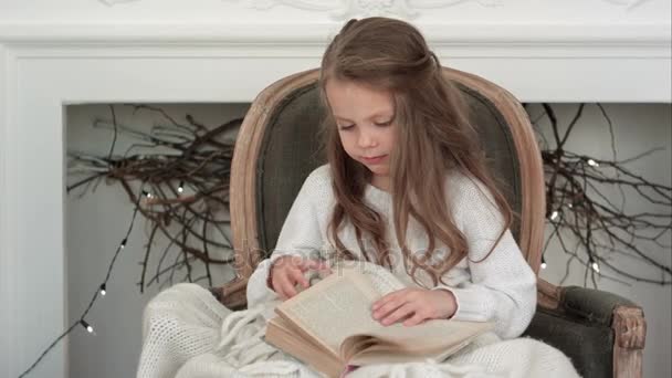 Linda niña sentada en una silla de Navidad y revisando un libro — Vídeos de Stock