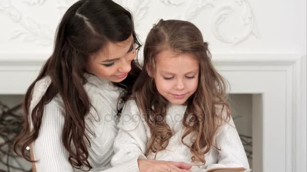 幸福的母亲和她漂亮的女儿在读一本书，在圣诞节前夕的白色毛衣 — 图库视频影像