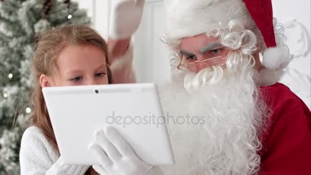 Санта Клаус делает рождественское селфи на планшете с милой маленькой девочкой, сидящей у него на коленях — стоковое видео