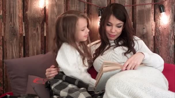 Fiatal anya, egy könyv feküdt a kanapén, és vele a kis lányom átölelve, karácsonyi díszített ház belső — Stock videók
