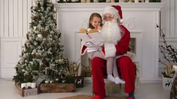 Санта-Клаус читает книгу с симпатичной девушкой возле елки — стоковое видео