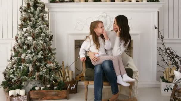 Joven madre hablando por teléfono con una hija ligeramente aburrida sentada en su regazo junto al árbol de Navidad — Vídeo de stock