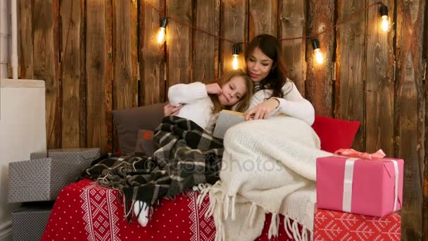 Bastante joven mamá leyendo un cuento de Navidad a su linda hija sentada en el sofá envuelta en mantas — Vídeo de stock