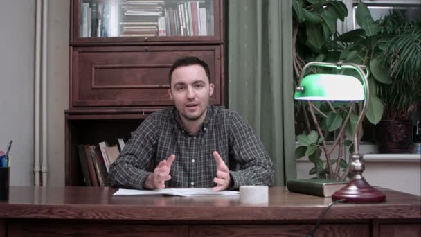 Ung man sitter vid skrivbord med papper och talar om hans betänkande i kameran — Stockvideo