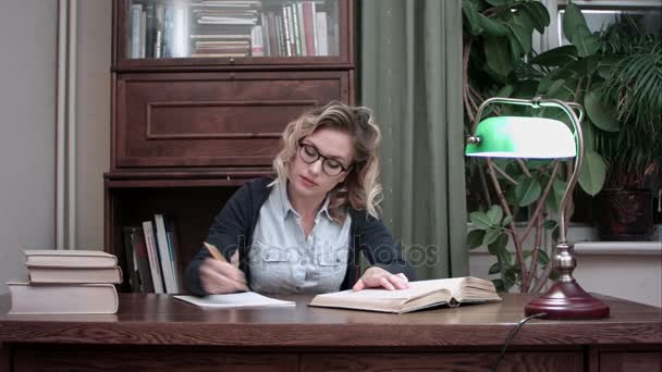 Joven estudiante de gafas sentado en una mesa y escribiendo notas de un libro — Vídeos de Stock