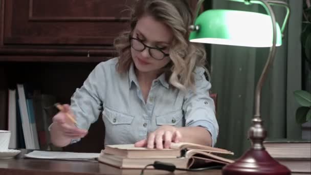 Estudiante ocupada con gafas trabajando en su informe en el escritorio con una lámpara verde, revisando libros y tomando café — Vídeo de stock