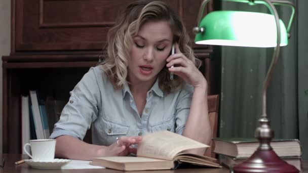 Young woman going through a book and having an angry phone conversation sitting at her desk — Stock Video