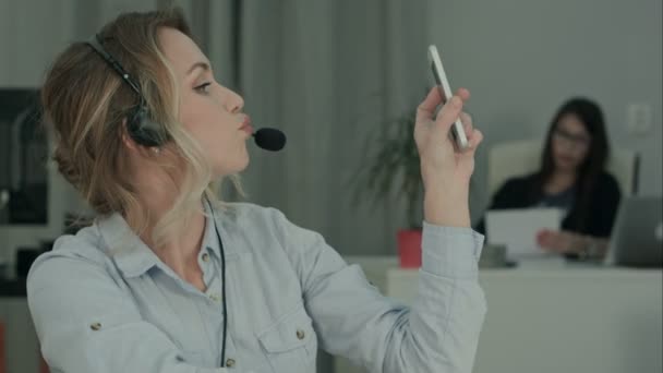Trabajador de oficina bastante joven en auriculares tomando selfies divertidos en el lugar de trabajo — Vídeos de Stock