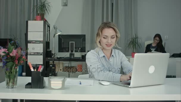 Улыбающаяся женщина печатает на ноутбуке и делает заметки за рабочим столом — стоковое видео