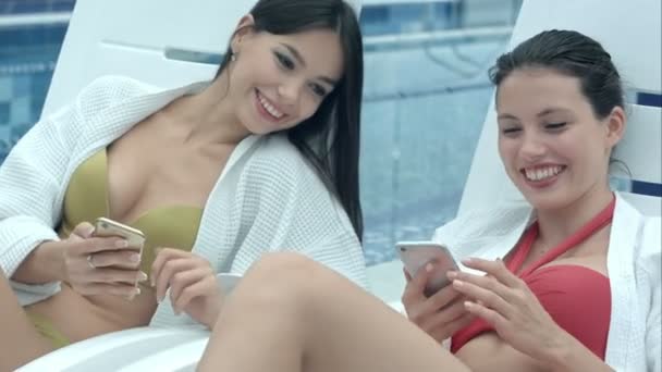 Duas mulheres bonitas felizes deitado na piscina chaise longues e mostrando uns aos outros fotos de seus telefones — Vídeo de Stock