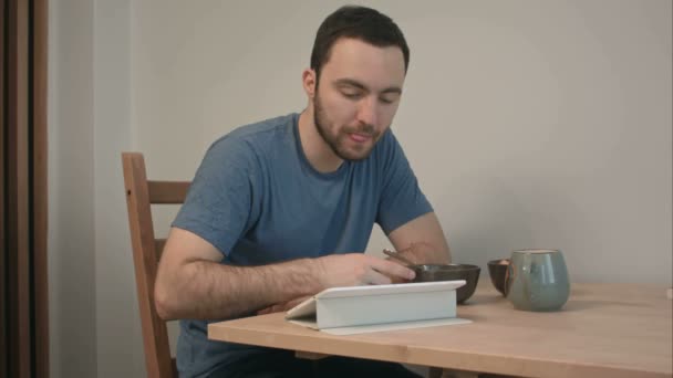 Молодой человек завтракает за чтением новостей на планшетном компьютере — стоковое видео