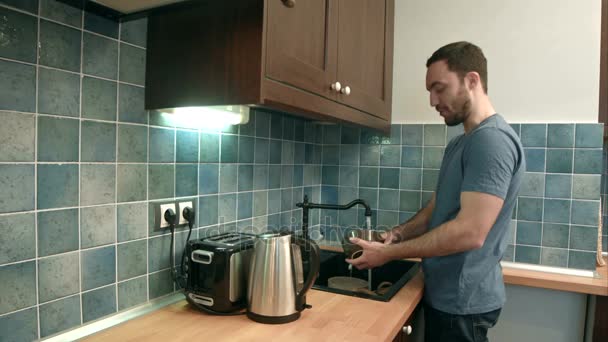 Улыбающийся молодой человек моет посуду и разговаривает с камерой — стоковое видео
