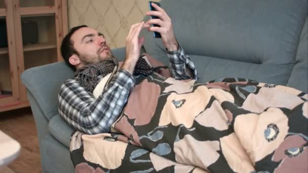 Больной молодой человек лежит на диване со своим телефоном — стоковое видео