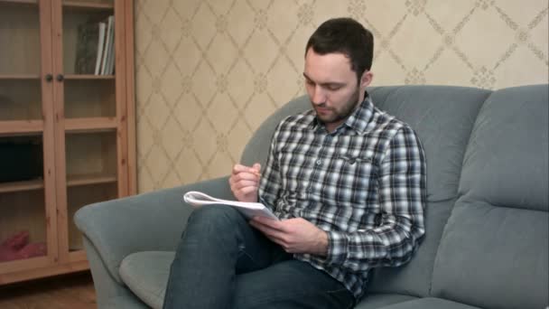 Молодой человек сидит на диване и делает заметки в учебнике. — стоковое видео