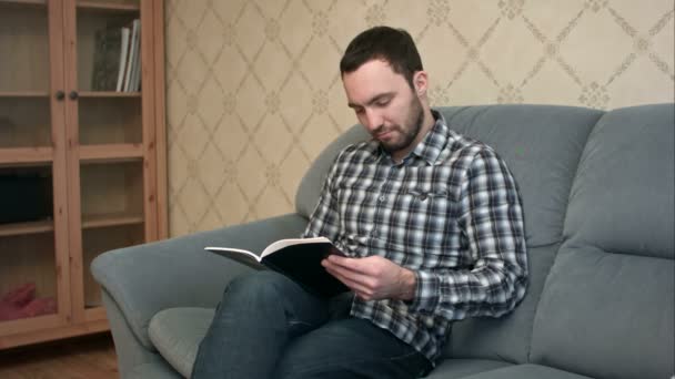 Konzentrierter junger Mann liest Buch auf dem Sofa — Stockvideo