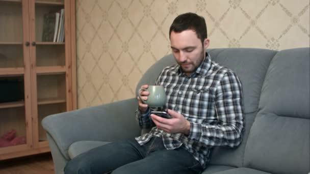 Young man with smartphone sitting on couch at home — Stock Video