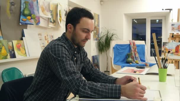 Manliga konststuderande att göra en skiss med en penna som sitter vid skrivbordet i studio — Stockvideo
