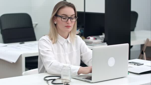 Концентрированная молодая женщина в очках, печатающая на ноутбуке у себя в офисе — стоковое видео