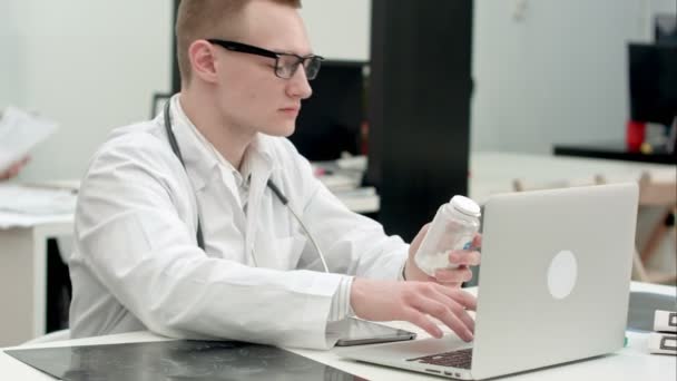 Médico masculino sosteniendo tarro de pastillas y mecanografiando prescripción en portátil — Vídeos de Stock