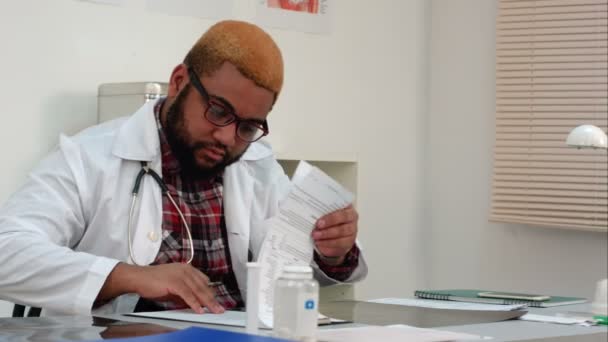 Afroamerican physician filling in medical forms and answering phone calls at his desk — Stock Video