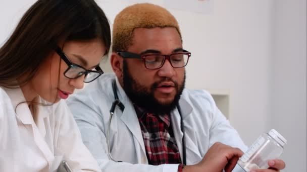 Афроамериканский врач держит банку с таблетками, пока медсестра делает заметки — стоковое видео