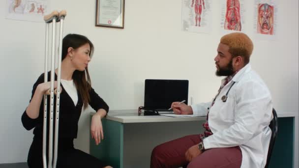 Female patient with crutches discussing her condition with doctor making notes — Stock Video