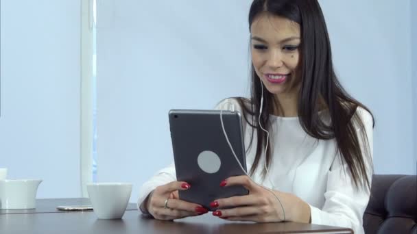 Femme souriante dans les écouteurs ayant un appel vidéo via une tablette numérique dans un café — Video