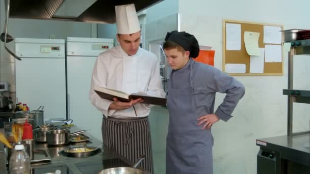 Jefe de cocina sosteniendo libro de cocina y explicando algo a su joven aprendiz — Vídeo de stock