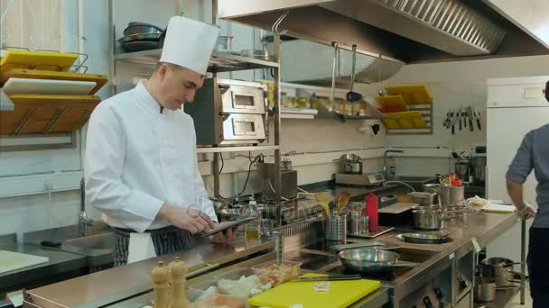 Hoofd koken met behulp van Tablet PC terwijl jonge assistent voorbereiding pan — Stockvideo