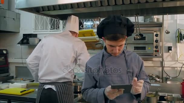 Ung laga trainee använda surfplatta samtidigt kock upptagen matlagning i köket — Stockvideo