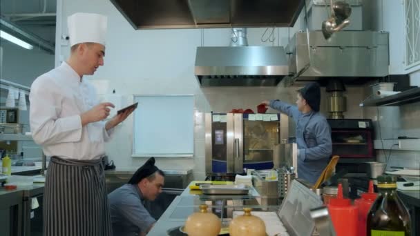 Chef sosteniendo la tableta discutiendo algo con un joven aprendiz mientras otro cocinero prepara sartenes — Vídeos de Stock