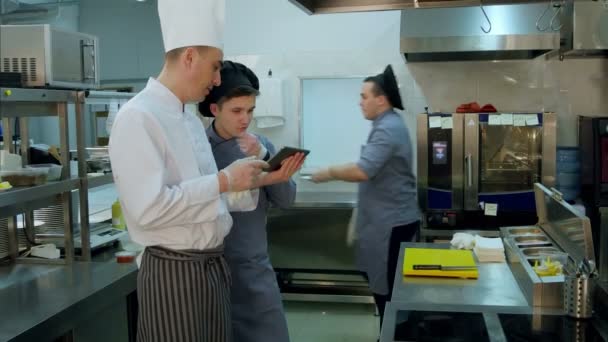 Шеф-повар объясняет что-то готовить стажёра в шляпе с помощью цифрового планшета — стоковое видео