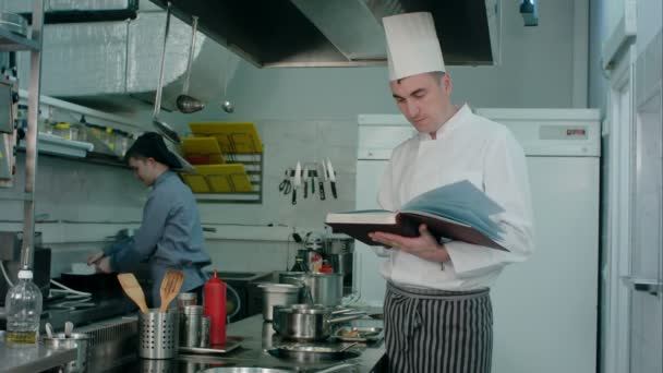Capo chef in cerca di ricetta in un libro, mentre il controllo sul suo tirocinante cuoco — Video Stock