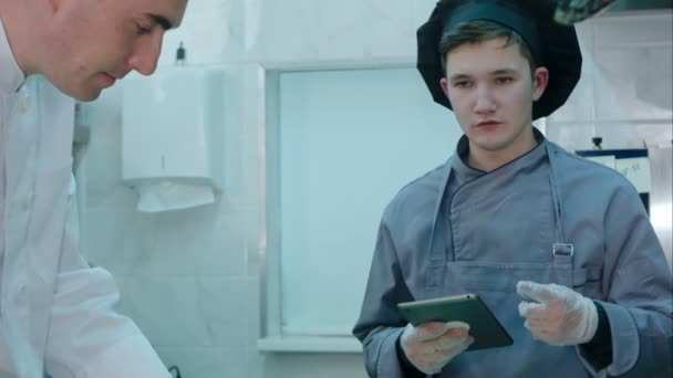 Jovem cozinheiro estagiário com tablet fazendo perguntas do chef sobre o processo de cozimento — Vídeo de Stock