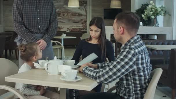 Familia joven con hija cenando en el restaurante. Padre llama al camarero para que su esposa ordene . — Vídeos de Stock