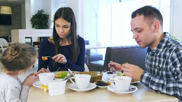 Jóvenes padres encantadores alimentando a su hija rubia ensalada de verduras en la cafetería . — Vídeo de stock