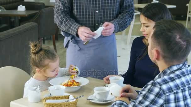 Menina engraçada brincando com seu brinquedo antes de comer seu jantar no café com seus pais. Empregado traz-lhe um plugue . — Vídeo de Stock