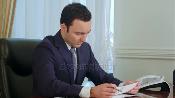 Молодой бизнесмен читает бумажки за столом в офисе и улыбается — стоковое видео